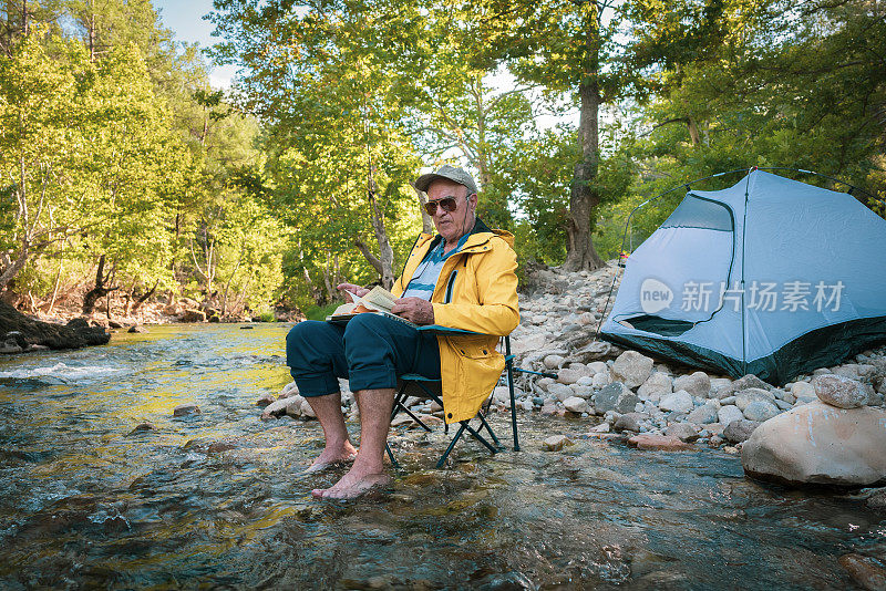 一位老人坐在森林小溪里的露营椅上看书