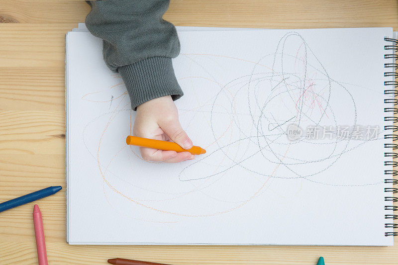一个幼儿用彩色蜡笔在木桌上的画册上画画。俯视图