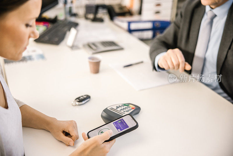一位女顾客在一家汽车经销店的室内办公桌旁，与一名汽车销售人员坐在一起，用信用卡上的近场通讯(NFC)支付了新车费用后，正在低头看智能手机