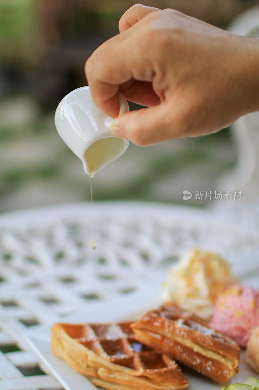 用手将枫糖浆浇在华夫饼和花园白色桌子上的苹果冰淇淋上。茶时间的情景在明朗的心情背景下。健康食物概念