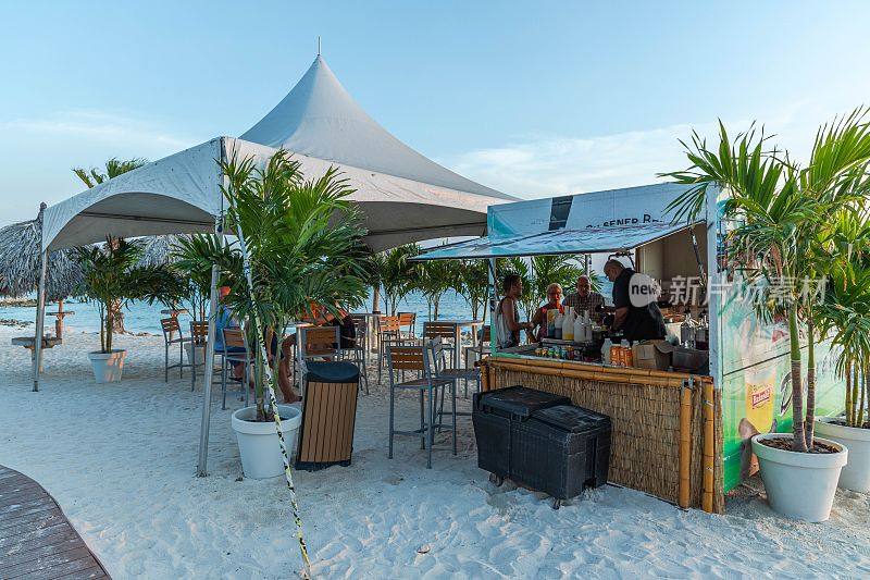 观看游客在户外酒吧排队喝酒。白色的沙滩和棕榈树在蓝天的背景下。