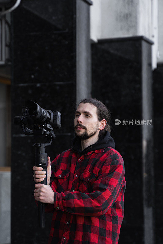 年轻的专业摄像师手持专业摄像机，安装在三轴框架稳定器上。专业设备有助于制作高质量的视频而不摇晃。一名身穿红衬衫的摄影师正在拍摄录像。