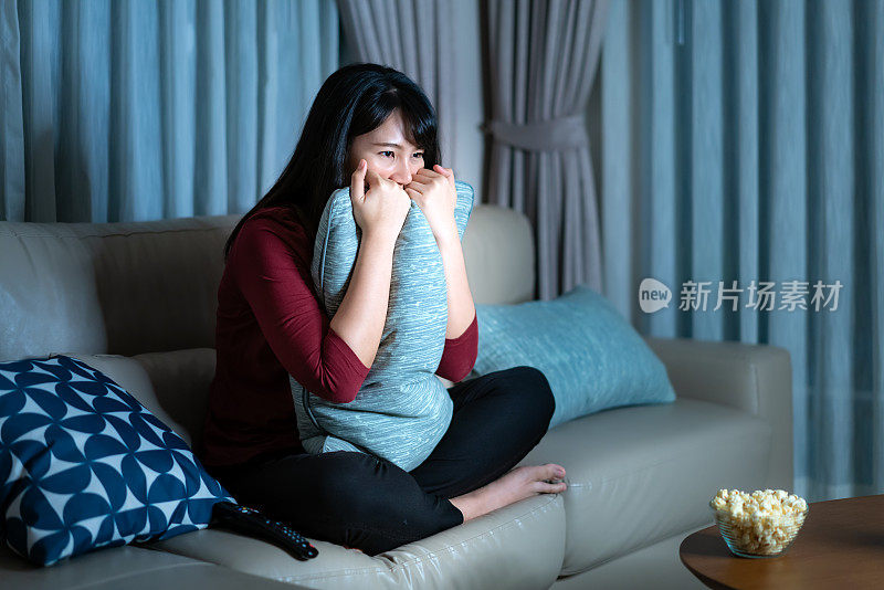 年轻的亚洲女人看电视悬疑电影或新闻，看起来震惊和兴奋深夜在家里客厅的沙发上吃爆米花。