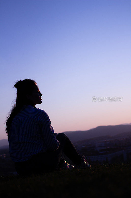 一个女人的黑色剪影坐在草山上俯瞰风景。