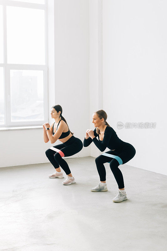 两名女子带着乳胶带微笑地在健身房训练。两名女孩一边做仰卧起坐，一边高兴地看着对方。