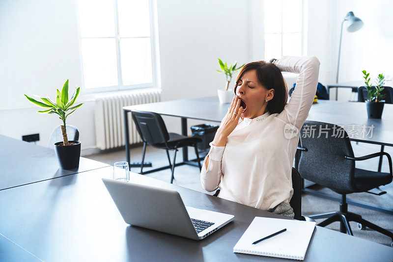 一位年轻的商务女性坐在现代办公桌旁，对着笔记本电脑打哈欠。