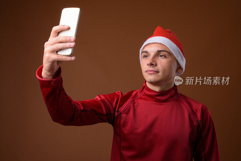 在彩色背景下，一名年轻帅气的男子穿着红色夹克，戴着圣诞帽，正在用手机拍照