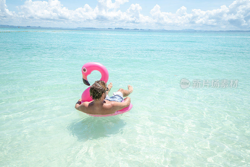 一个年轻人在田园般的海滩上放松，充气火烈鸟在清澈的海水中玩耍在泰国的岛屿。人们旅行奢侈，乐趣和酷的态度概念