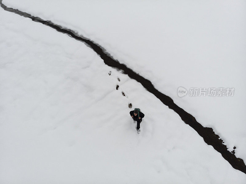 成年男子徒步行走在白雪覆盖的田野上，旁边是小水流。鸟瞰图。