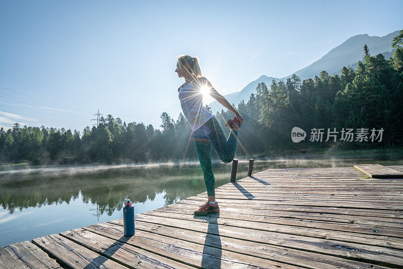 年轻女子伸展身体晨跑，日出在美丽宁静的湖面上，女性站在码头上水上