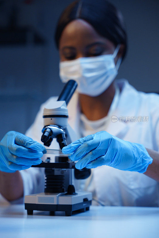 一位非裔美国医生坐在实验室的办公桌前，用显微镜载玻片和显微镜工作