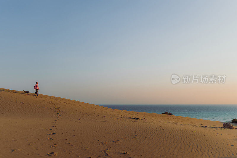 在土耳其，一个带着狗的女人在海边的沙丘上凝视着风景优美的日落