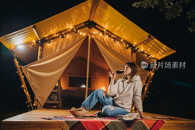 微笑的女性自由职业者喝着酒，看书坐在舒适的野营帐篷在秋天的晚上。户外度假度假的豪华露营帐篷。生活方式的概念
