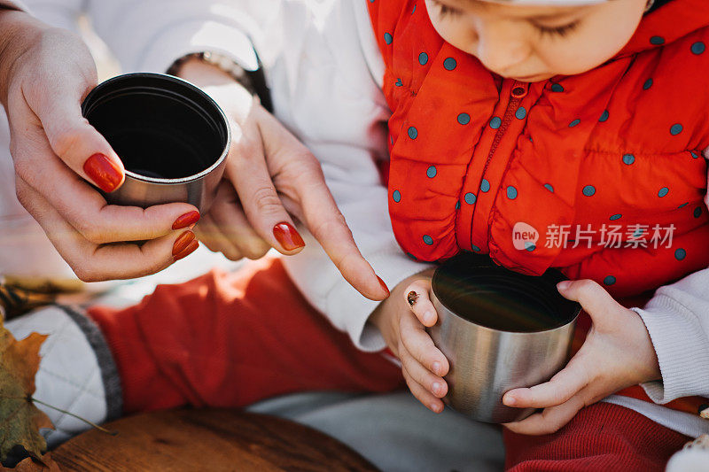 幸福的家庭母亲和蹒跚学步的小女儿在秋天野餐，用红色的保温瓶和杯子