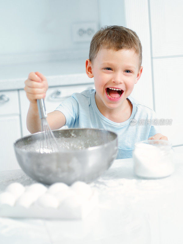 孩子在做饭。一个白人男孩坐在厨房的桌子旁，用手打蛋器在杯子里揉面。开朗积极的孩子做派，笑。年轻的厨师或厨师。孩子的肖像。