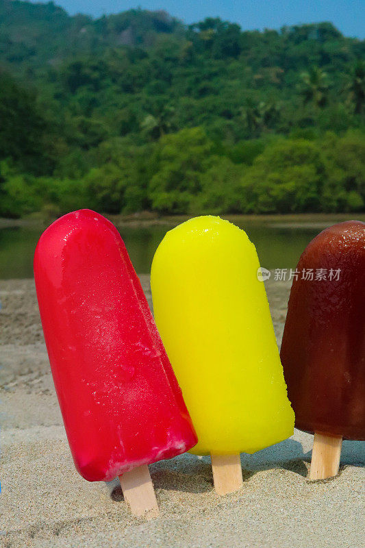 图片:三排冰棒，木棒棒卡在沙滩上，退潮时在海边的水边有红色，黄色和棕色的冰棒，草莓，柠檬和可乐口味的冰棒，以海岸为背景，重点在前景，复制空间
