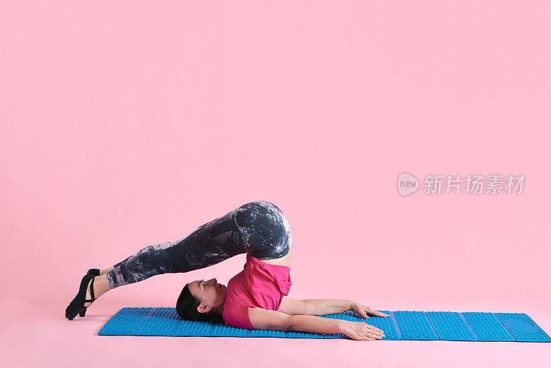 灵活的身体。成熟的女性在健身matt上训练，在粉红色的工作室背景下伸展身体