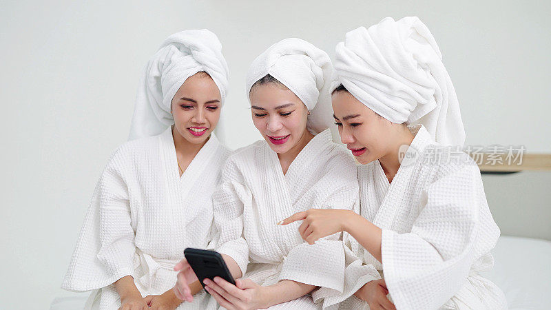 三位女性朋友穿着浴袍和毛巾，坐在床上玩智能手机。友谊的概念