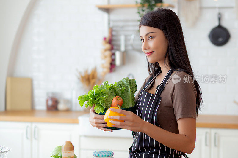 快乐的年轻亚洲妇女在家里的厨房里拿着蔬菜篮子准备烹饪沙拉蔬菜食品，妇女站着提着新鲜的蔬菜篮子，生活方式的概念。