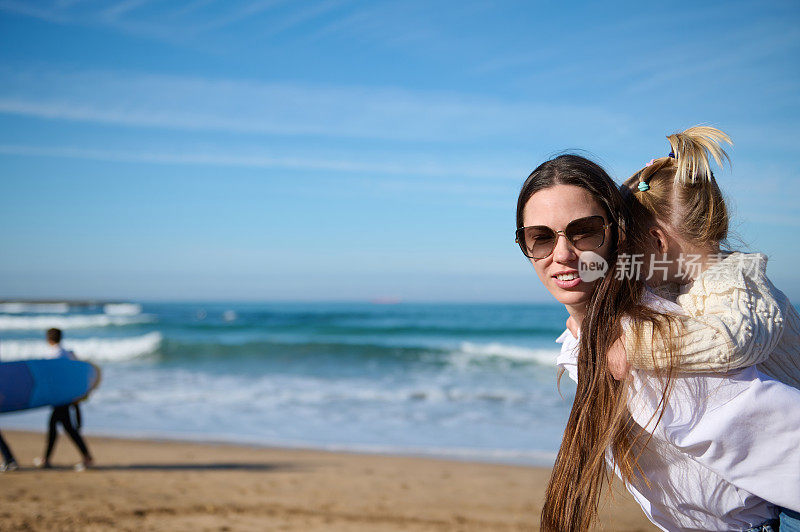 快乐的母亲和可爱的女儿在一起玩得很开心，在沙滩上散步，面对着不认识的人练习冲浪