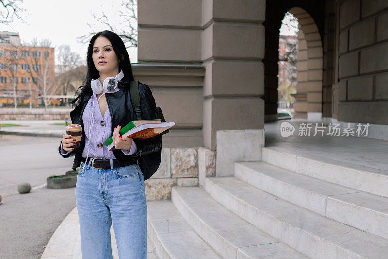 年轻漂亮的女大学生背着双肩包站在大学的楼梯上。她戴着耳机，穿着皮夹克，等待着大学课程的开始。