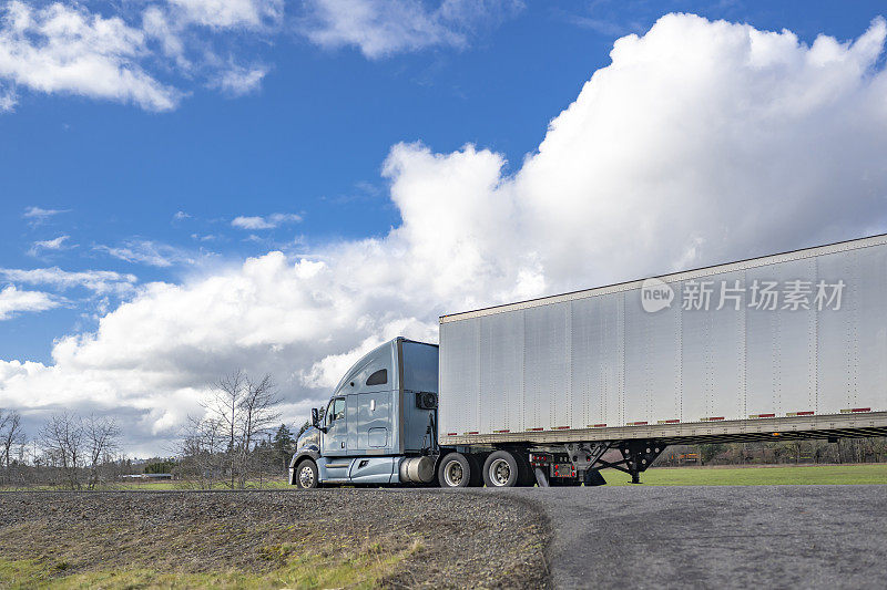 浅蓝色大卡车工业半挂车运输货物的干货车半挂车行驶在当地狭窄的道路上，背景是多云的天空