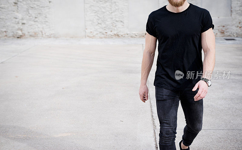 年轻的肌肉男穿着黑色t恤和牛仔裤走在市区。模糊的背景。Hotizontal模型。