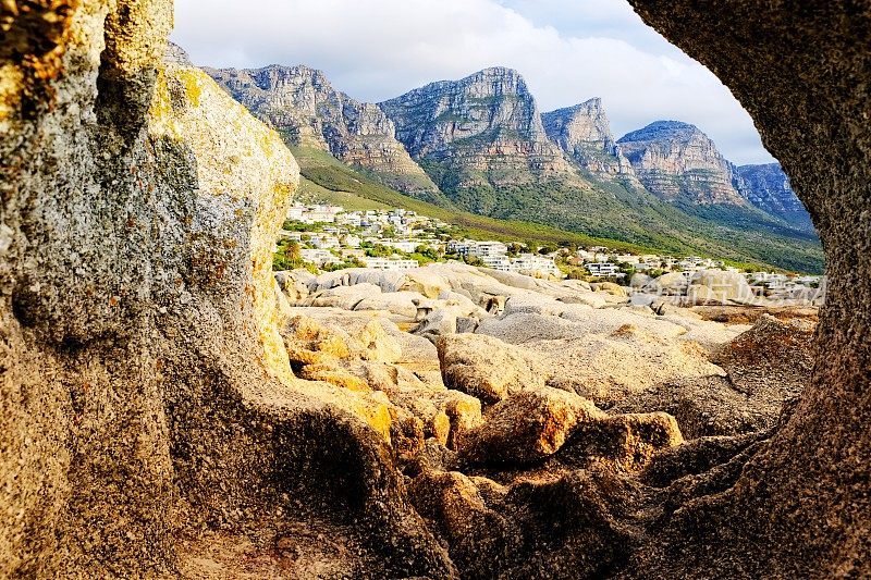 透过南非坎普斯湾海滩的一块岩石，可以看到十二使徒山脉脚下的房屋和公寓楼