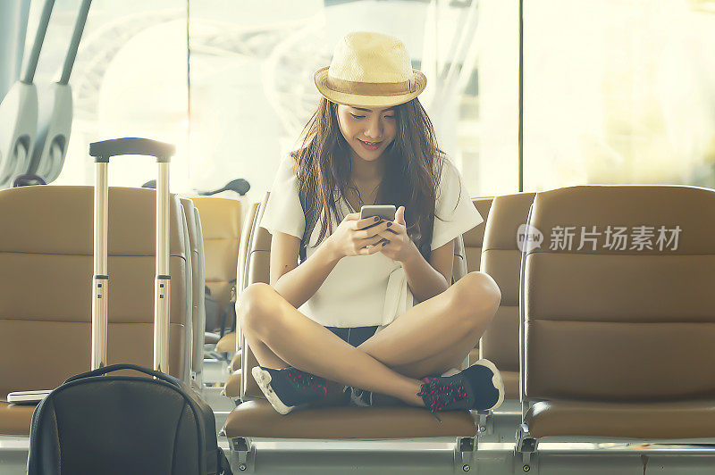 年轻女子在机场用智能手机等待飞机。她推着旅行箱手推车坐在机场候机室的候机室里。旅行的概念。