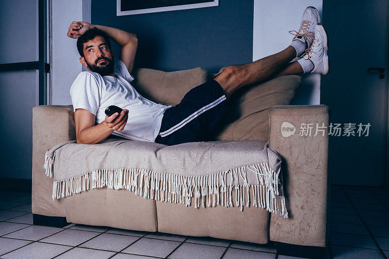 男人穿着运动服躺在沙发上看电视。懒惰的概念