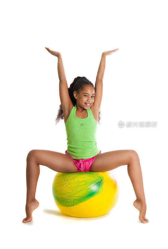 一个漂亮的黑皮肤女孩在一个黄色的球上做体操。女孩举起双手，微笑着，快乐着，