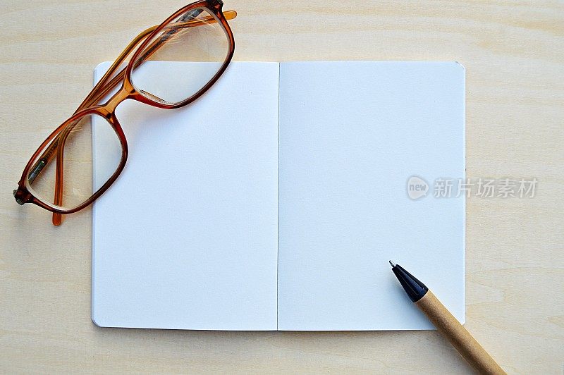 一张水平的照片，一张白色的素色记事本，连同一支棕色和黑色的钢笔和眼镜，放在一个木制的米色水平背景上，很有美感。