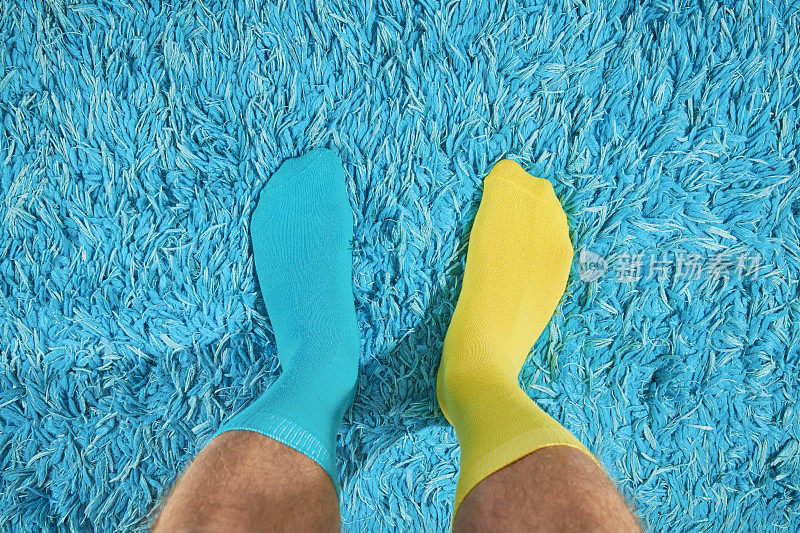 POV的观点，男人的脚穿着奇怪的袜子，蓝色和黄色对抗一个明亮的蓝色地毯。