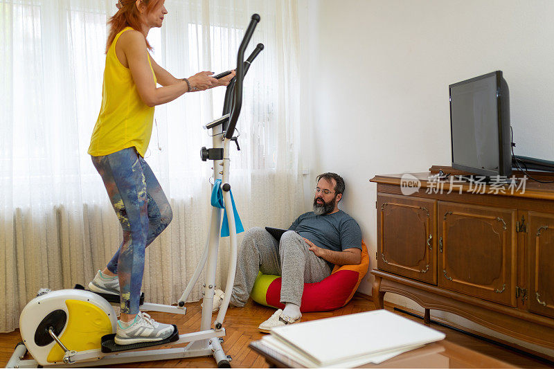 女人在椭圆机上锻炼，而她的伴侣在客厅里使用平板电脑