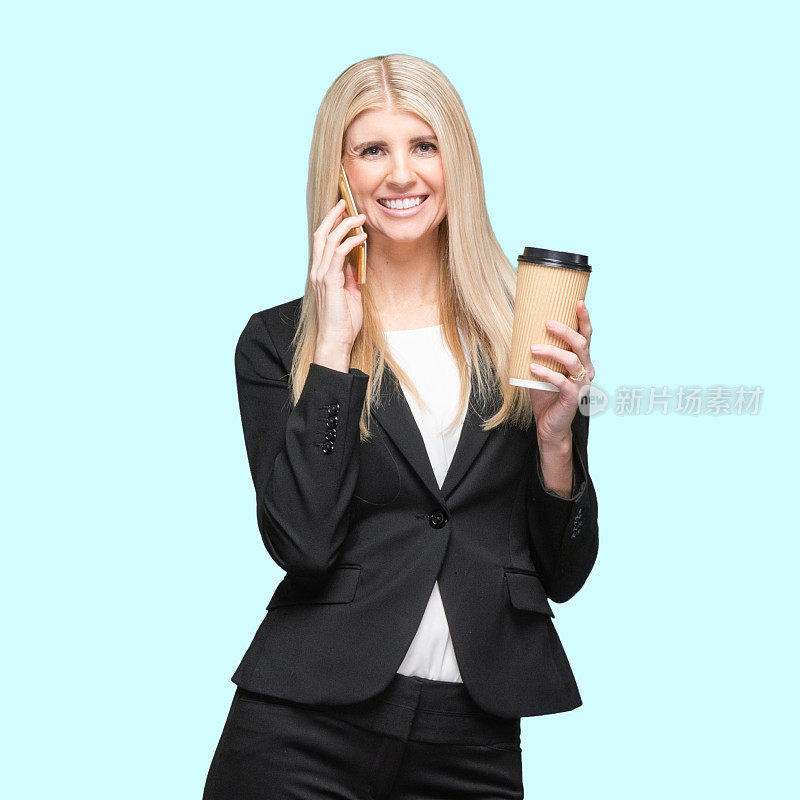 白人女商人站在有色背景前，穿着夹克，拿着咖啡杯，使用手机