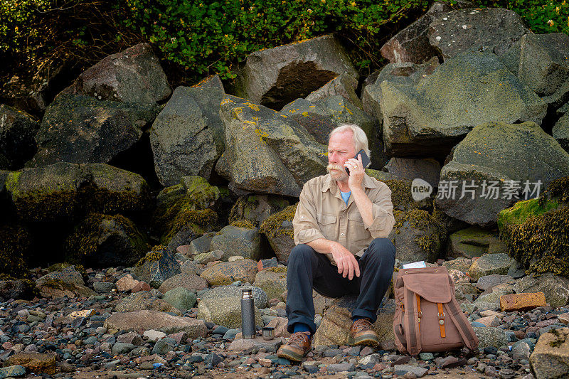 一位白发苍苍的老人坐在岸边的岩石上玩手机