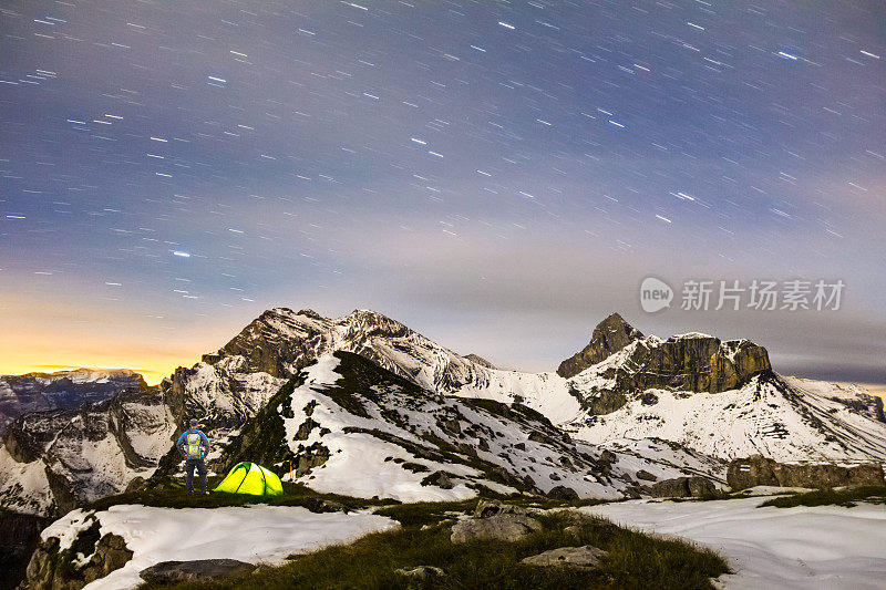 一个人站在帐篷旁边，在雪山星空下。瑞士阿尔卑斯山脉。