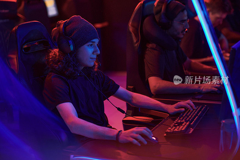 年轻的卷发玩家头戴帽子，头戴耳机，手持电脑与团队进行在线游戏互动