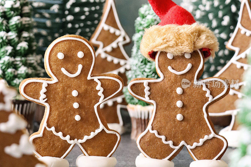 两个自制的姜饼人的特写图像，装饰着白色的皇家糖霜，在池塘上滑冰，一个戴着红色的圣诞帽，在白雪覆盖的圣诞森林场景中，模型杉木和纸杯蛋糕圣诞树，雪白的浅蓝色背景