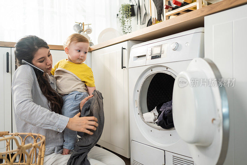 白人忙碌的母亲在厨房做家务和小男孩蹒跚学步。美丽的妈妈用电话上班，把衣服放到洗衣机里陪儿子在屋里玩。Family-housekeeping。