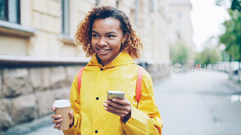 快乐的非裔美国妇女正在使用智能手机触屏，微笑着走在美丽的城市户外，喝着外带咖啡。现代的生活方式和沟通理念。