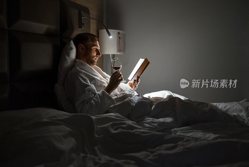 中年男子放松地躺在床上看书，拿着一杯红酒，床头灯开着。晚上放松，爱好，自由时间的概念。成年的概念。