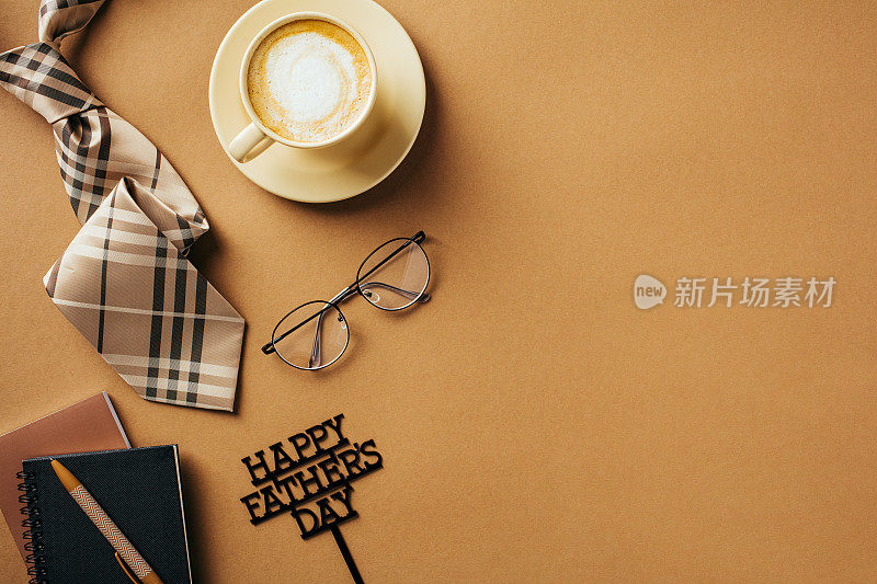 父亲节快乐的概念。平铺咖啡杯，领带，眼镜，纸质笔记本，在棕色背景上写上父亲节快乐。