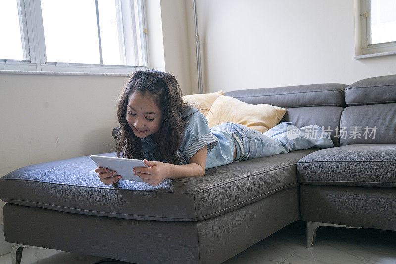 年轻女孩在家里的沙发上愉快地使用着数码平板电脑