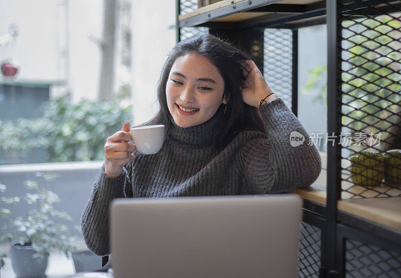 一名年轻的亚洲女子在咖啡馆边喝咖啡边摆弄电脑