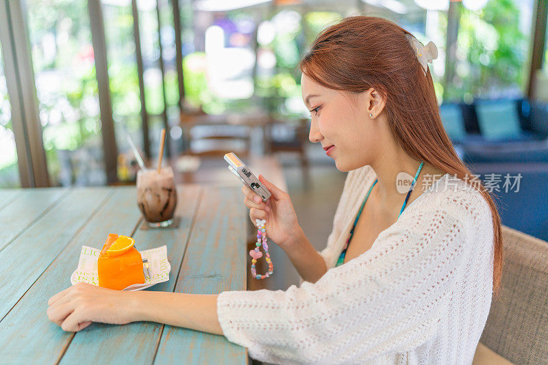 年轻的亚洲女子在海滩咖啡馆喝冰拿铁时，正在拍一块橘子蛋糕。女性数字游牧民穿着沙滩装在度假氛围中拍摄美食照片。穿着沙滩装的亚洲女人在咖啡馆里放松。