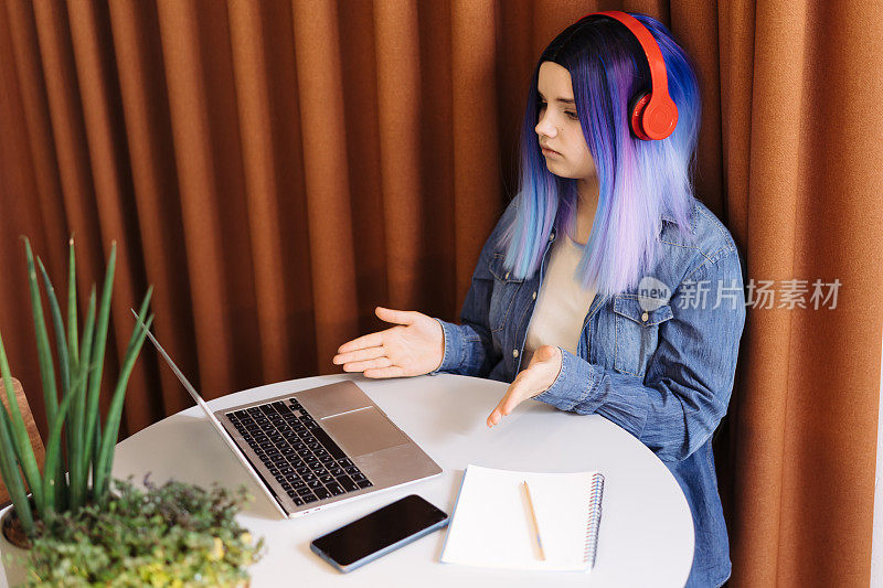 迷人的年轻拉丁裔女孩的肖像，蓝头发，蓝色牛仔衬衫坐在咖啡馆里，用笔记本电脑进行在线视频通话。本空间
