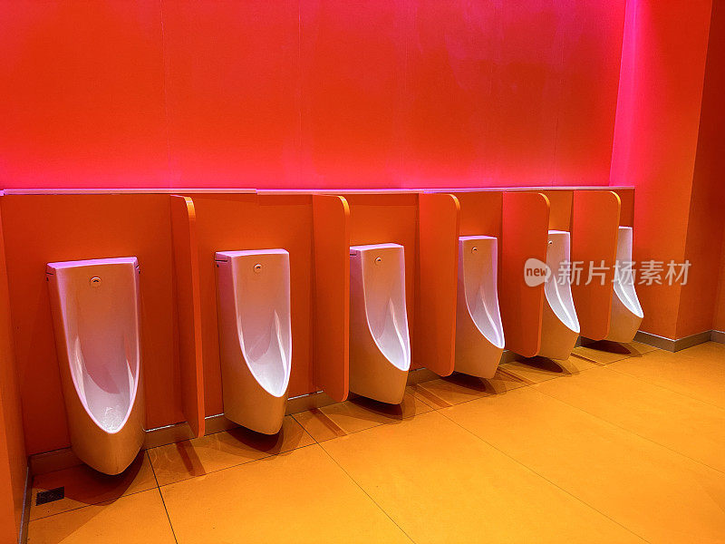 公共厕所里一排现代的白色陶瓷小便池，红色和橙色照明墙壁上的壁挂式碗，木制的隐私屏风隔板，背景飞溅，干净卫生的小便池，墙内有排水管和排水系统