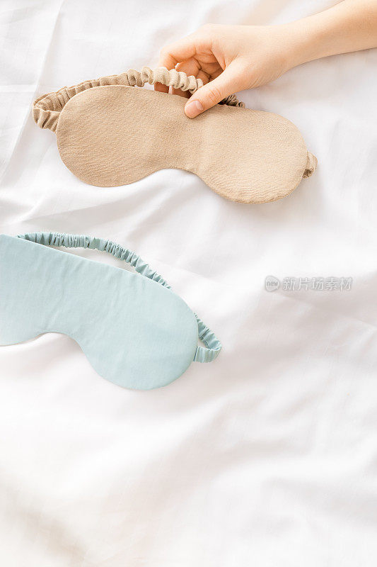 合上两个睡眠用的眼罩，米色和蓝色放在白色的被褥上，女人手拿一个，尽量平躺。俯视图睡眠面膜，最佳睡眠者，舒适放松。好好休息的概念。