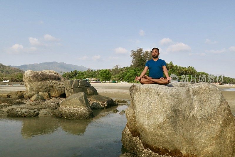 印度男子练习莲花姿势(莲花瑜伽)瑜伽位置在岩石，水的边缘，帕洛伦海滩，果阿，印度